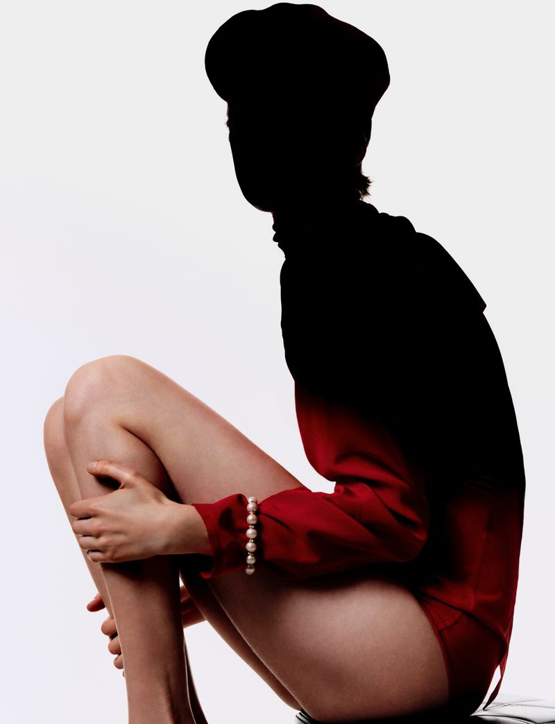 Seated woman in black cap and red bodysuit, partially silhouetted, pearl bracelet on wrist, against a plain white studio backdrop.