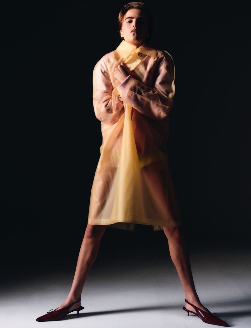 Fashion photo of a blonde adult male in a transparent yellow coat, posing in a dark studio with low-heeled, maroon pumps.