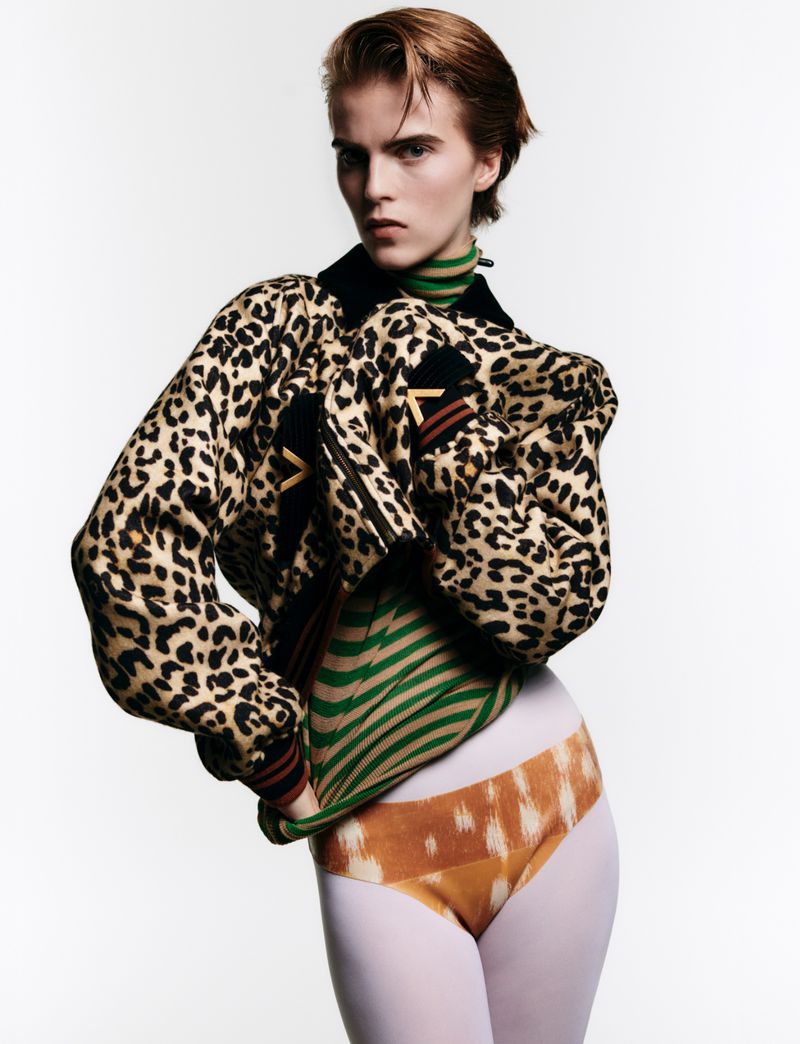 A fashion portrait of a young adult posing in a printed jacket with patterned top and bottoms against a bright white background.