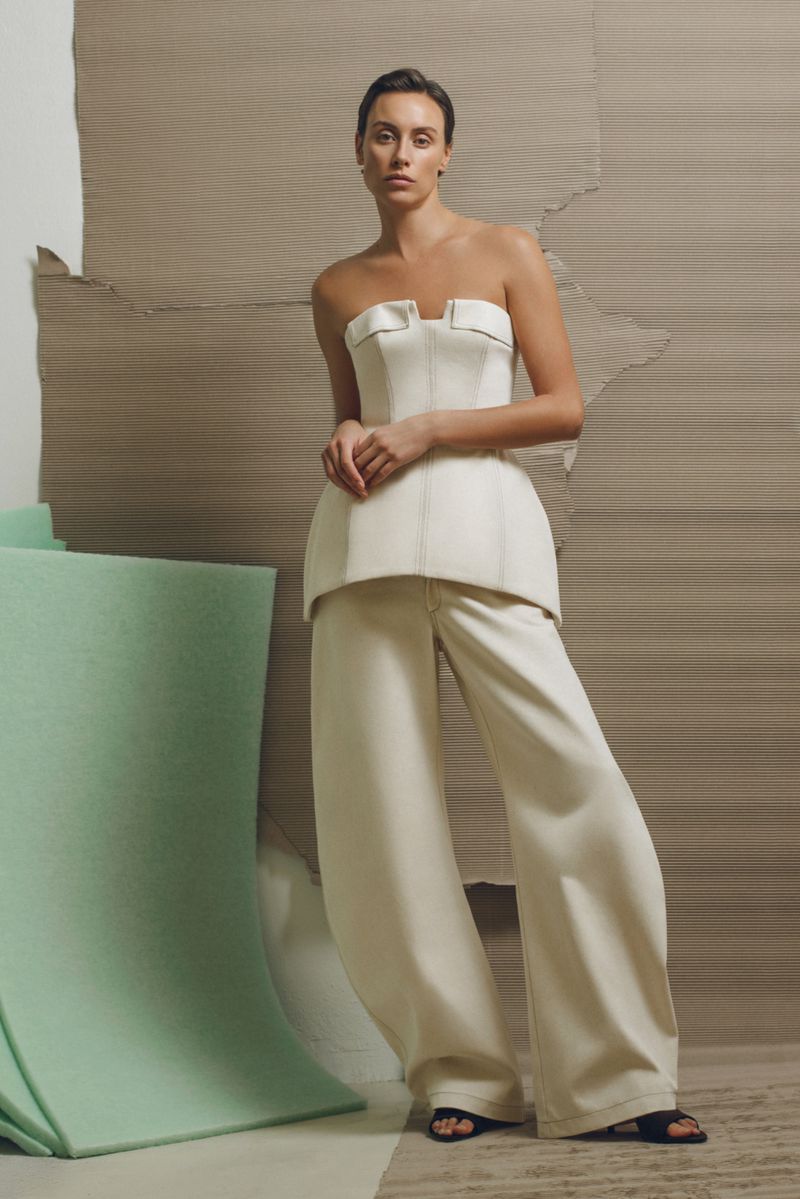 A woman in a cream-colored corset top and matching wide-legged pants poses against a tan background with a mint-green architectural element.