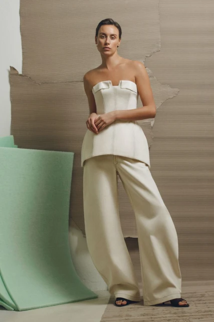 A woman in a cream-colored corset top and matching wide-legged pants poses against a tan background with a mint-green architectural element.