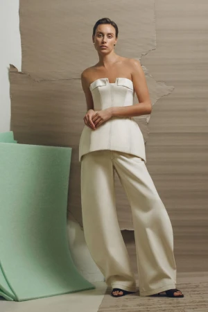 A woman in a cream-colored corset top and matching wide-legged pants poses against a tan background with a mint-green architectural element.