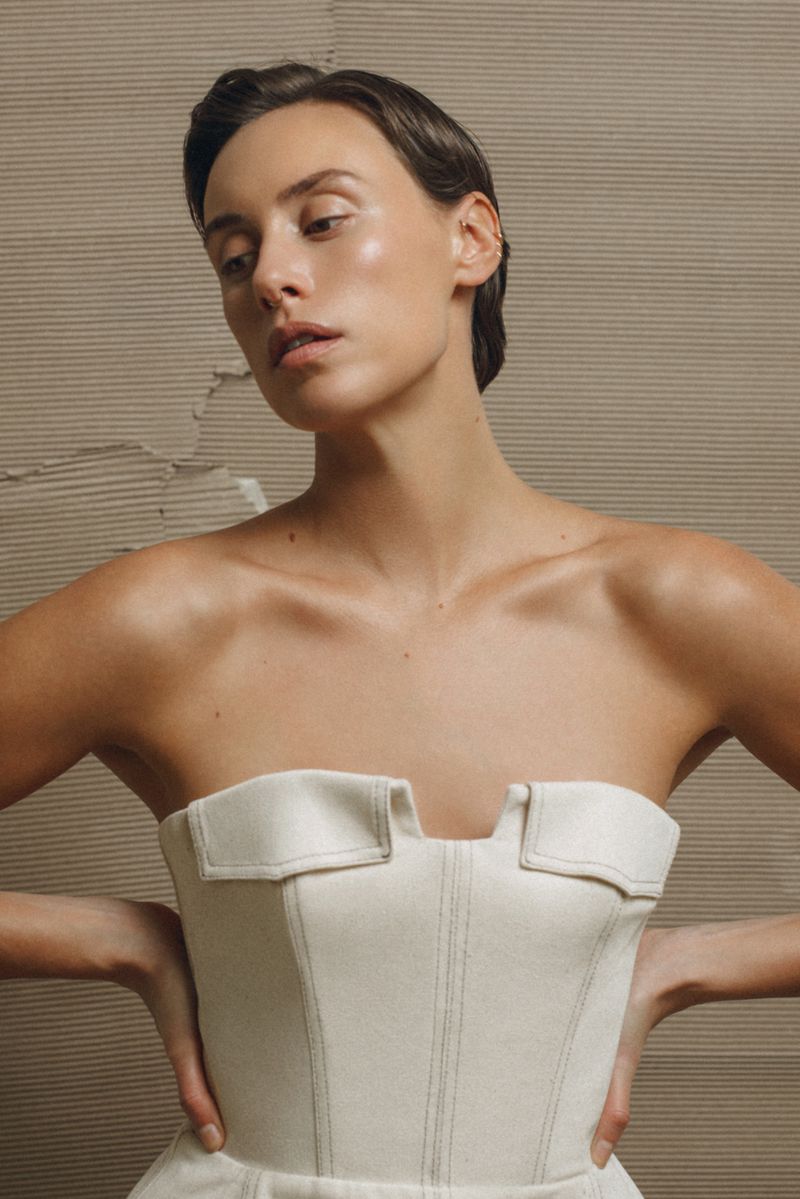 A woman with short dark hair poses in a strapless beige corset dress against a textured, neutral-toned backdrop in a studio setting.