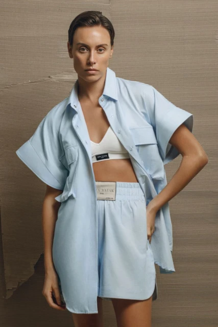 Woman in light blue shorts and open shirt, posing in front of a textured wall, fashion shoot, bright studio lighting, medium shot.