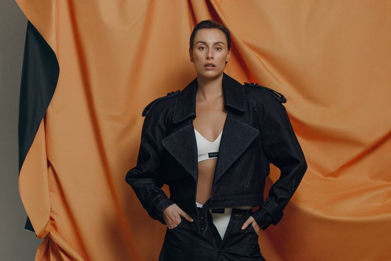 A woman poses in a dark blue denim jacket and off-white crop top against an orange and gray backdrop in a studio setting.