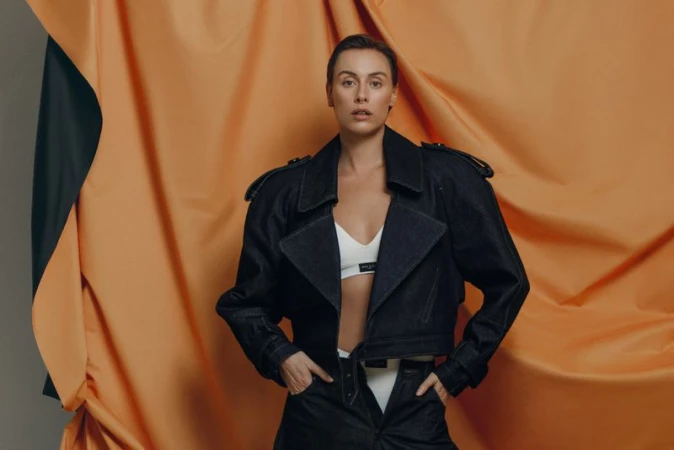 A woman poses in a dark blue denim jacket and off-white crop top against an orange and gray backdrop in a studio setting.