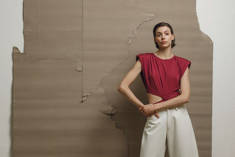 Fashion shot of a young adult woman in a red top and white pants posing in front of a textured cardboard backdrop in a studio setting.