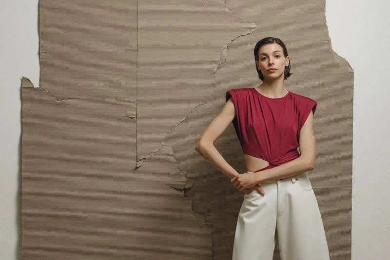 Fashion shot of a young adult woman in a red top and white pants posing in front of a textured cardboard backdrop in a studio setting.