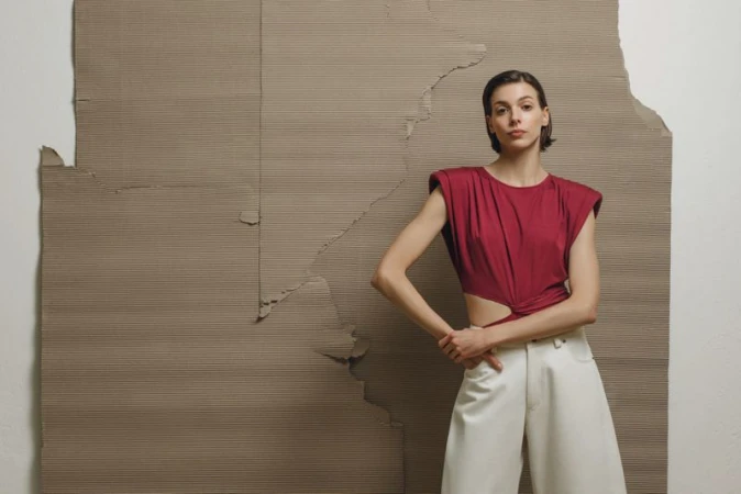 Fashion shot of a young adult woman in a red top and white pants posing in front of a textured cardboard backdrop in a studio setting.