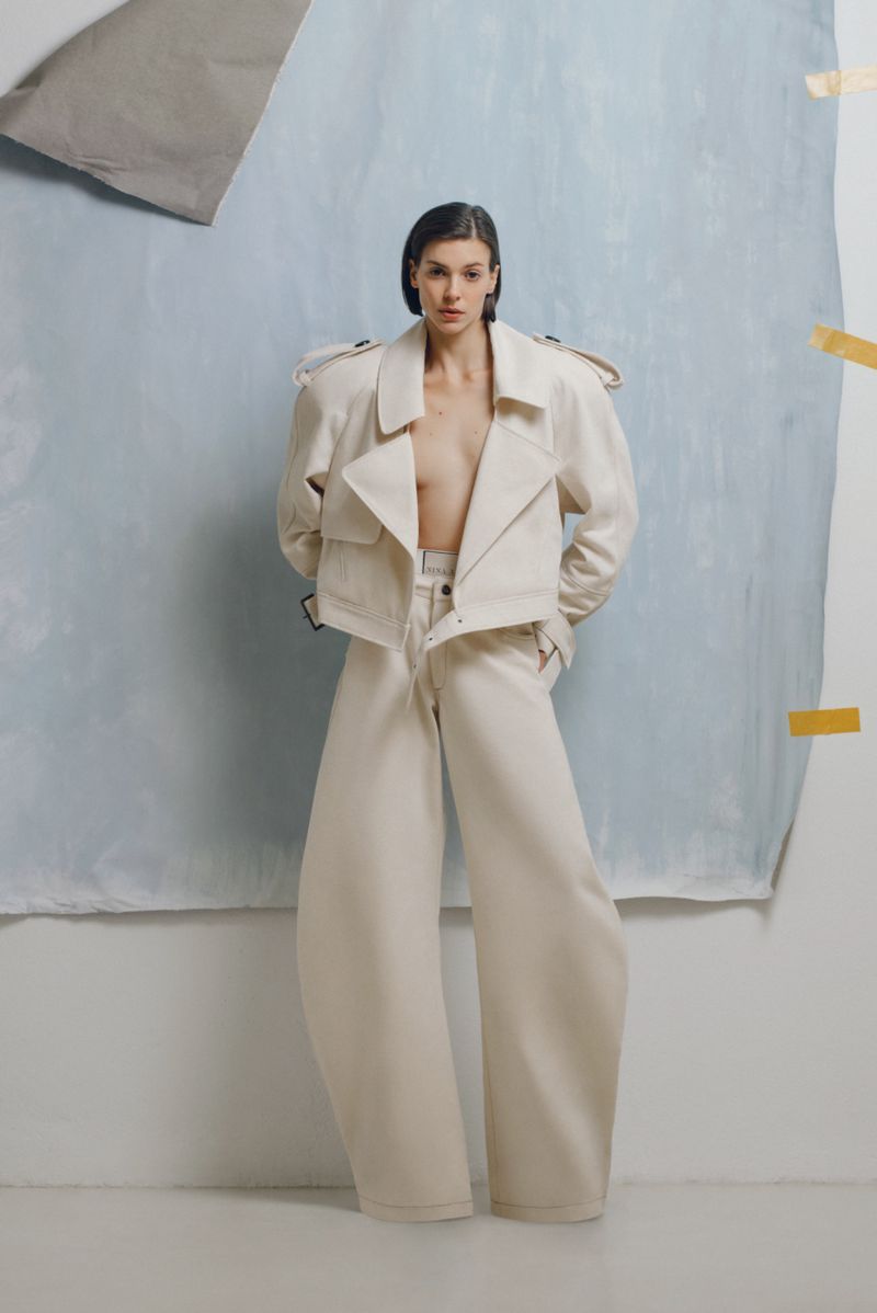 Fashion shot of a woman posing in a white jacket and pants against a painted backdrop in a studio setting with soft lighting.
