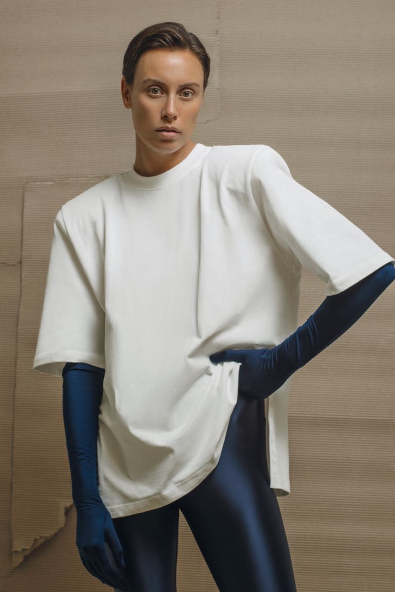 Adult female fashion model poses in a photo studio wearing a white t-shirt, blue gloves, and blue leggings against a neutral backdrop.