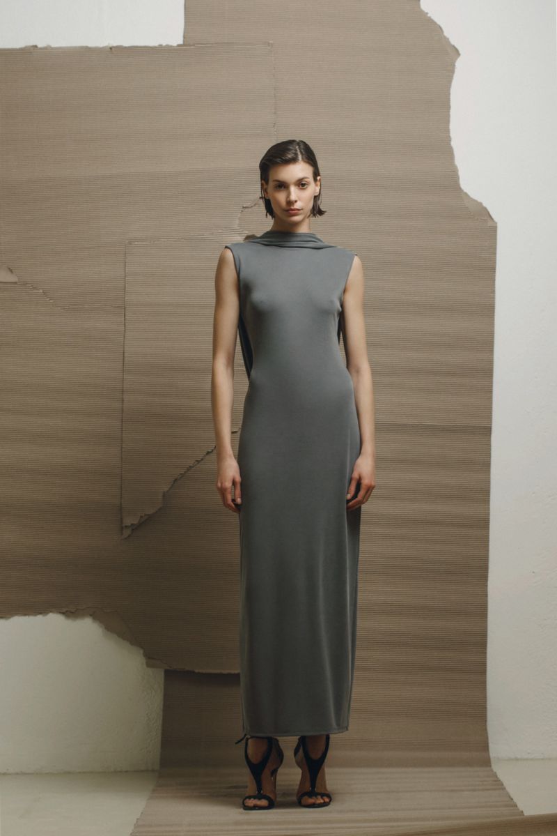 Full-length shot of a woman posing in a grey dress with black high-heeled sandals in front of a neutral backdrop in a studio.
