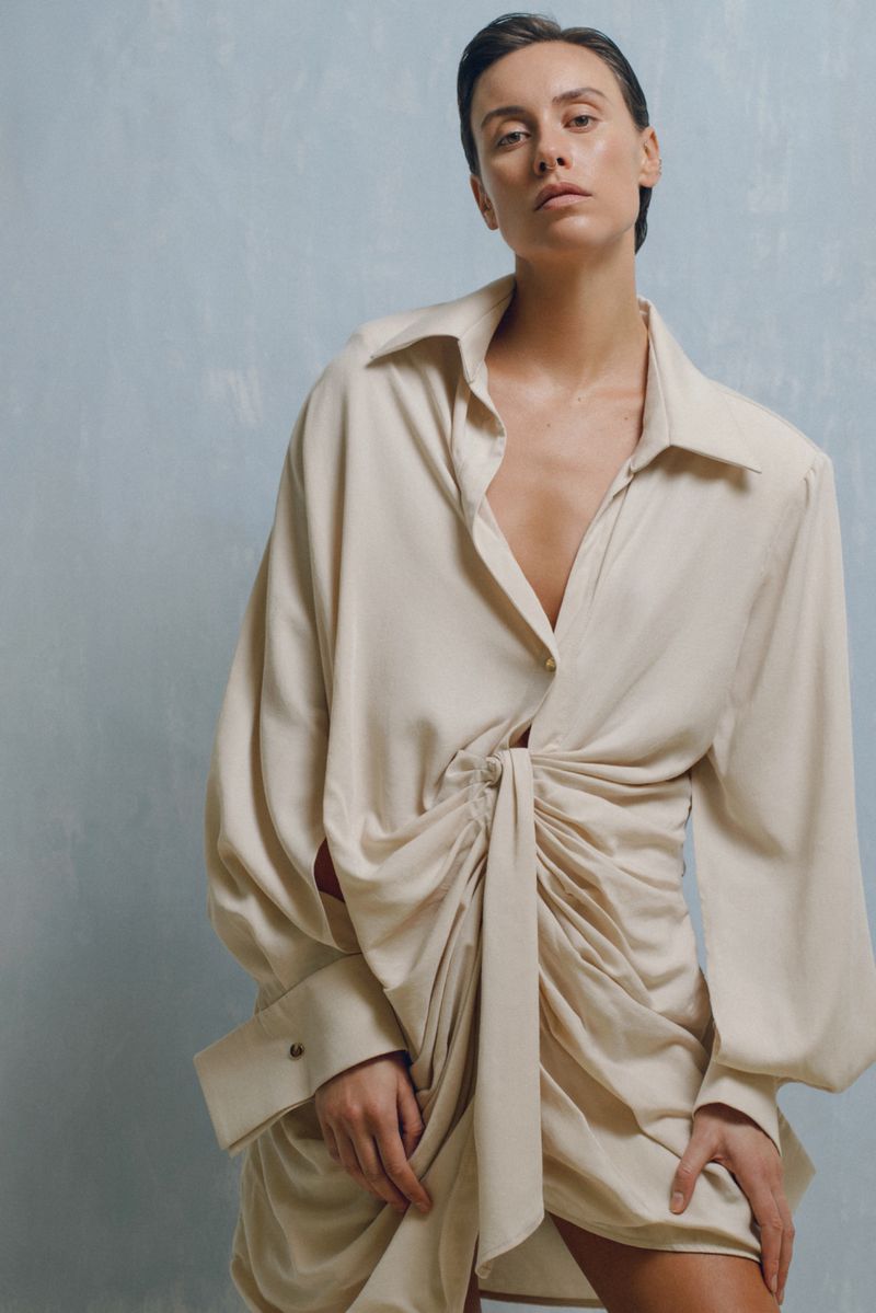 Woman poses in a beige, draped mini-dress against a textured blue background. Fashion studio shot with soft lighting and a neutral palette.