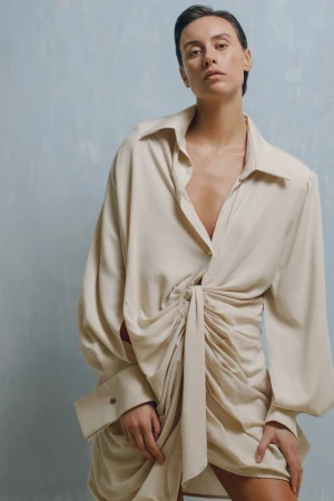 Woman poses in a beige, draped mini-dress against a textured blue background. Fashion studio shot with soft lighting and a neutral palette.