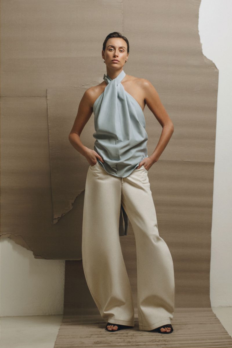 Woman in halter top and wide-leg trousers poses against neutral backdrop in fashion editorial, showcasing minimalist style.
