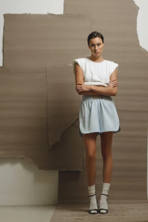 Model standing with arms crossed wearing a white top and light blue shorts against a layered cardboard backdrop