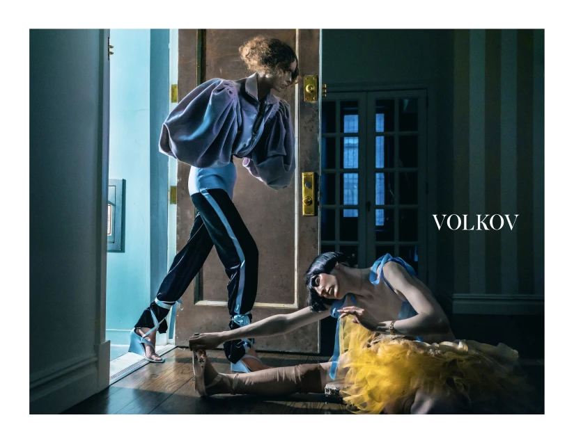 Two fashion models pose indoors in a dimly lit room. One stands by an open door in a mauve top and the other sits on the floor in a yellow tutu.