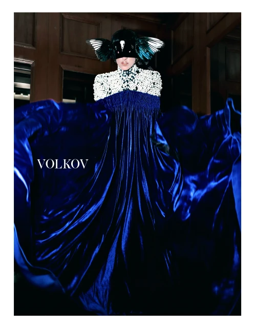 Person wearing a beaded dress and winged mask stands in front of a dark, paneled wall with a dark blue flowing fabric overlay.