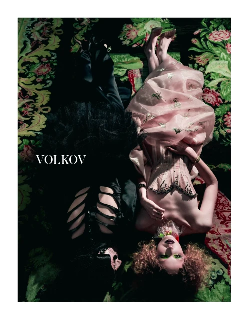 Fashion editorial featuring a man in black and a woman in pink lying on a floral patterned surface in a dramatic and moody setting.