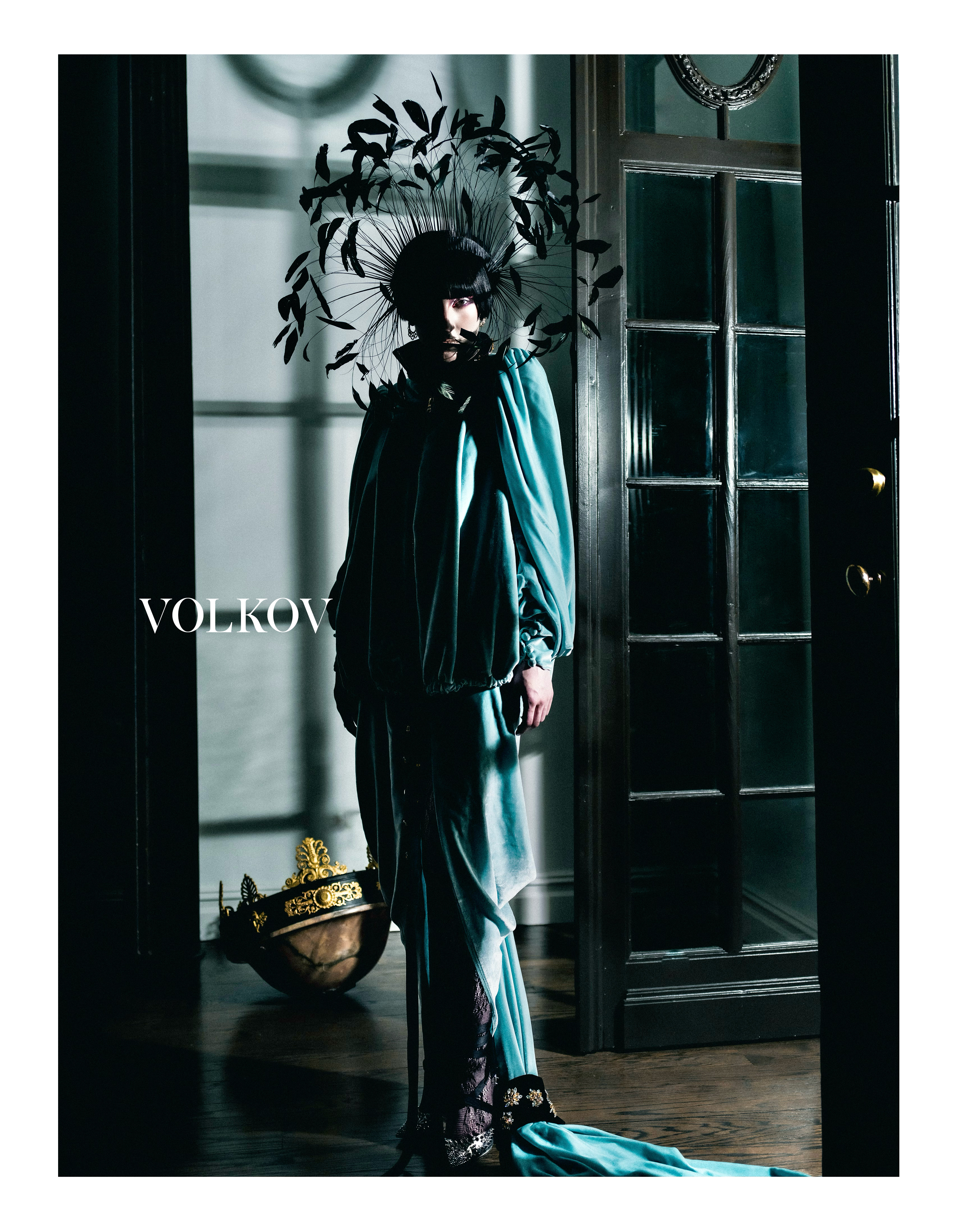 Fashion portrait of a person in a teal gown and black feathered headpiece standing in a dark, ornate hallway with wood floors.