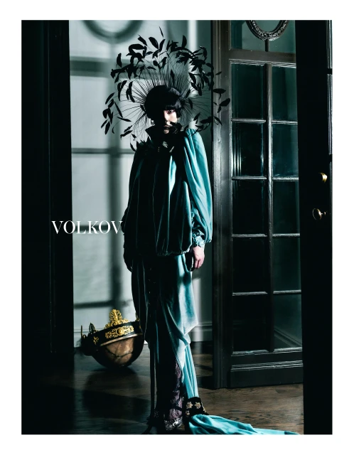 Fashion portrait of a person in a teal gown and black feathered headpiece standing in a dark, ornate hallway with wood floors.