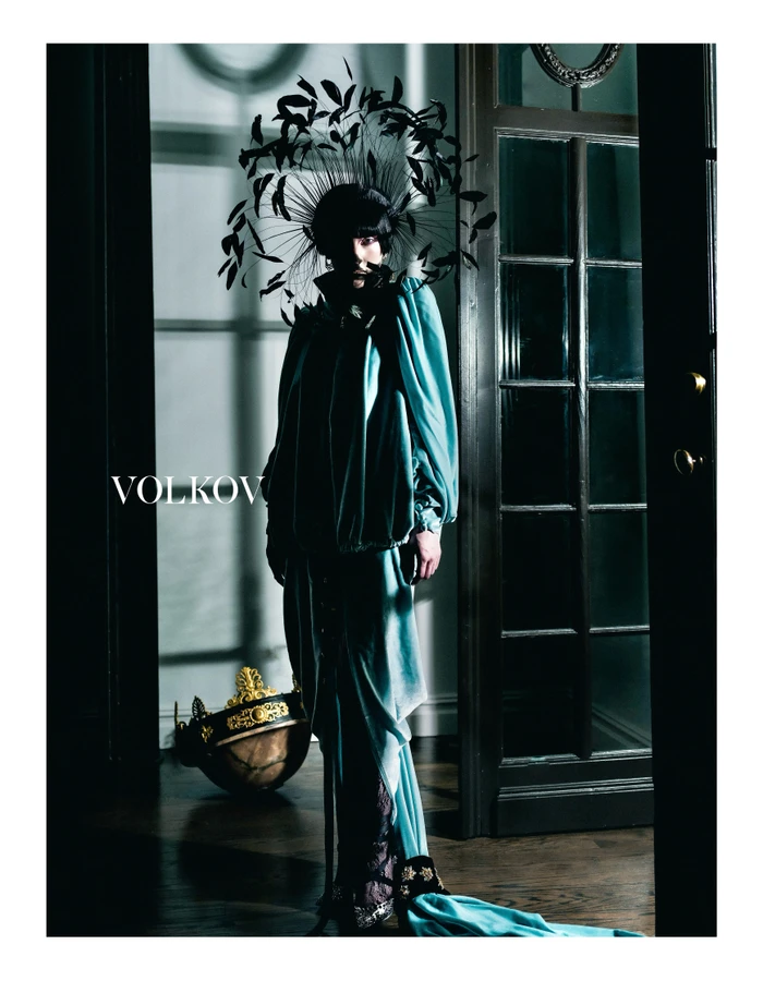 Model in teal velvet outfit with elaborate feathered headpiece standing in moody interior for Volkov FW25