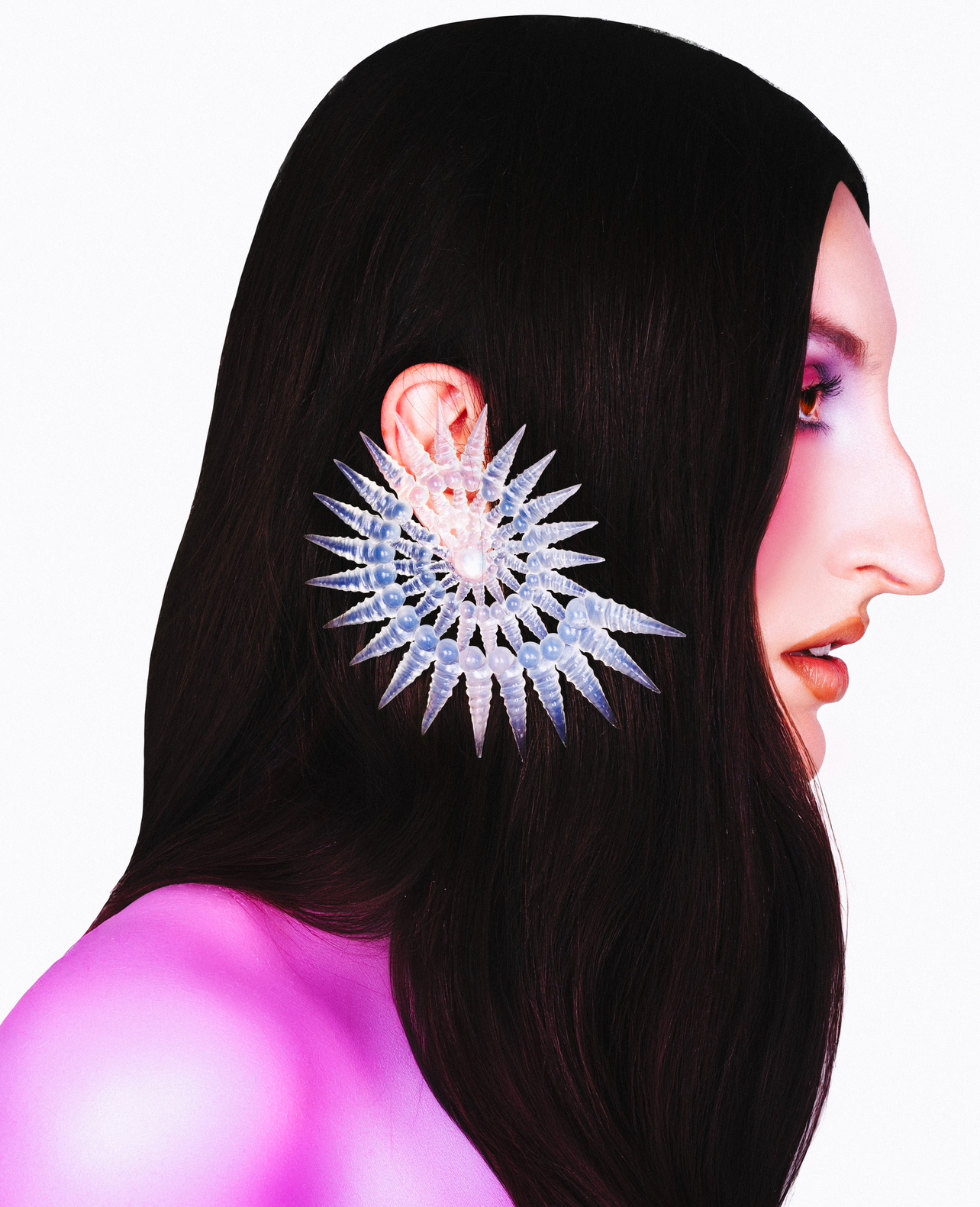 Profile of a woman with dark hair and dramatic makeup wearing an elaborate, translucent, spiked earring, against a white background.