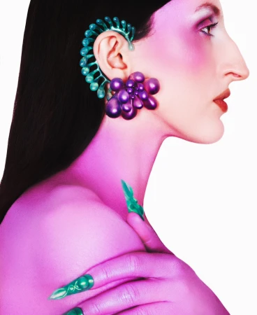 A side-profile studio shot of a woman with unique teal and purple sculptural earrings and green nail art on a white background.