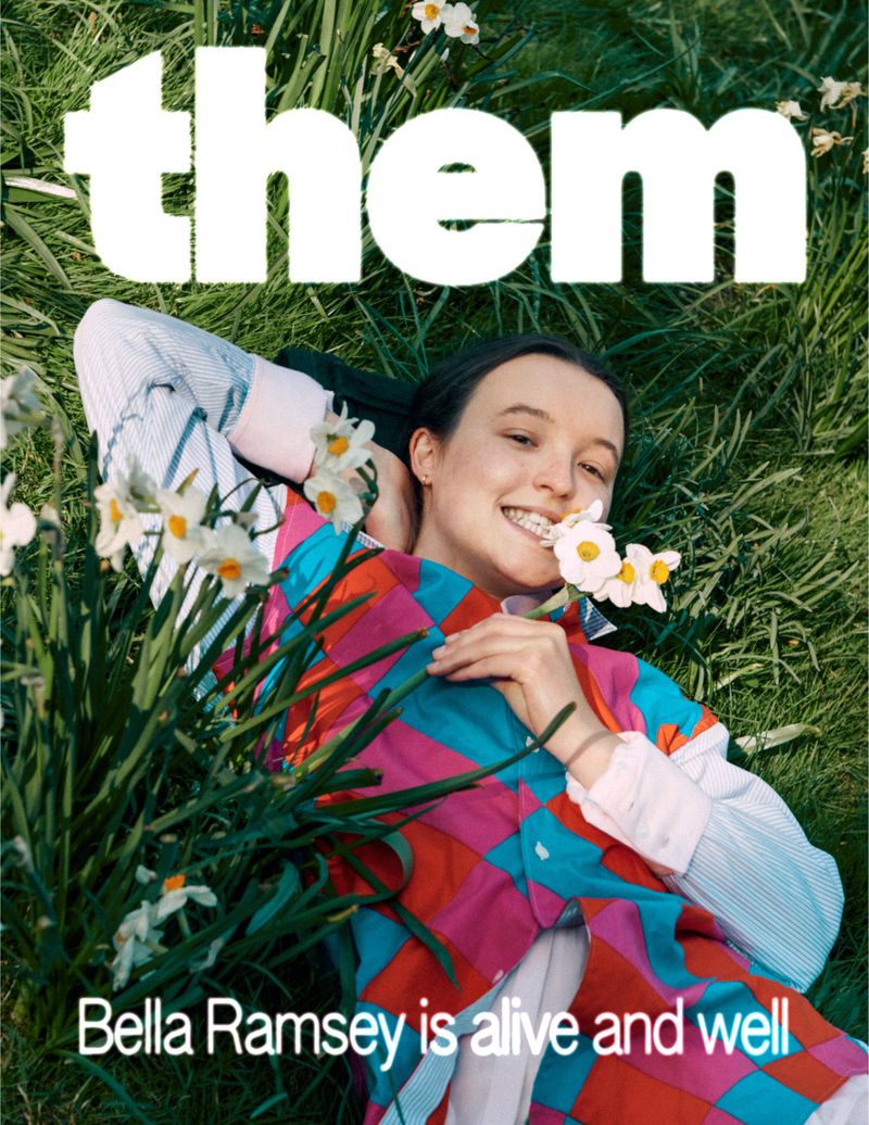 Bella Ramsey lies in grass with white flowers, smiling in a colorful shirt, as the cover for 'them' magazine with white text.