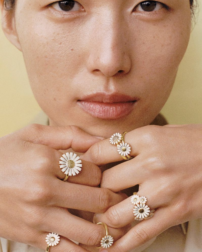 Close-up of an Asian woman's face and hands, showcasing multiple gold rings with daisy-shaped designs on her fingers. Beauty shot.