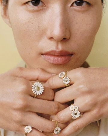 Close-up of an Asian woman's face and hands, showcasing multiple gold rings with daisy-shaped designs on her fingers. Beauty shot.