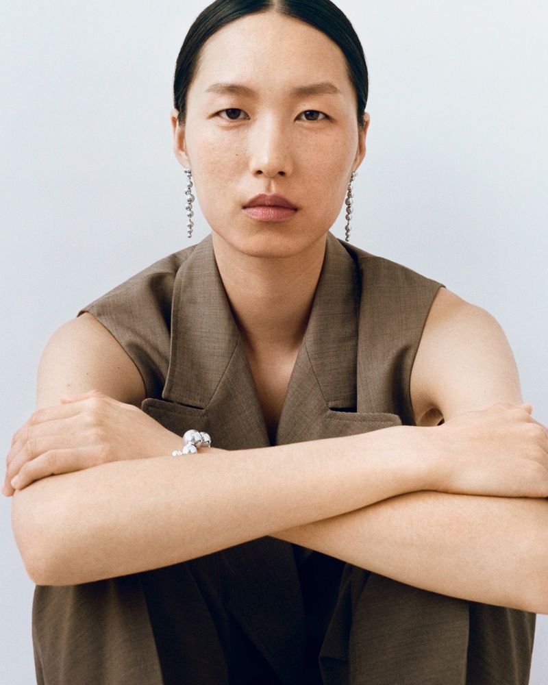 A stylish Asian woman in a brown sleeveless suit, seated indoors, accessorized with a silver bracelet and dangling earrings.