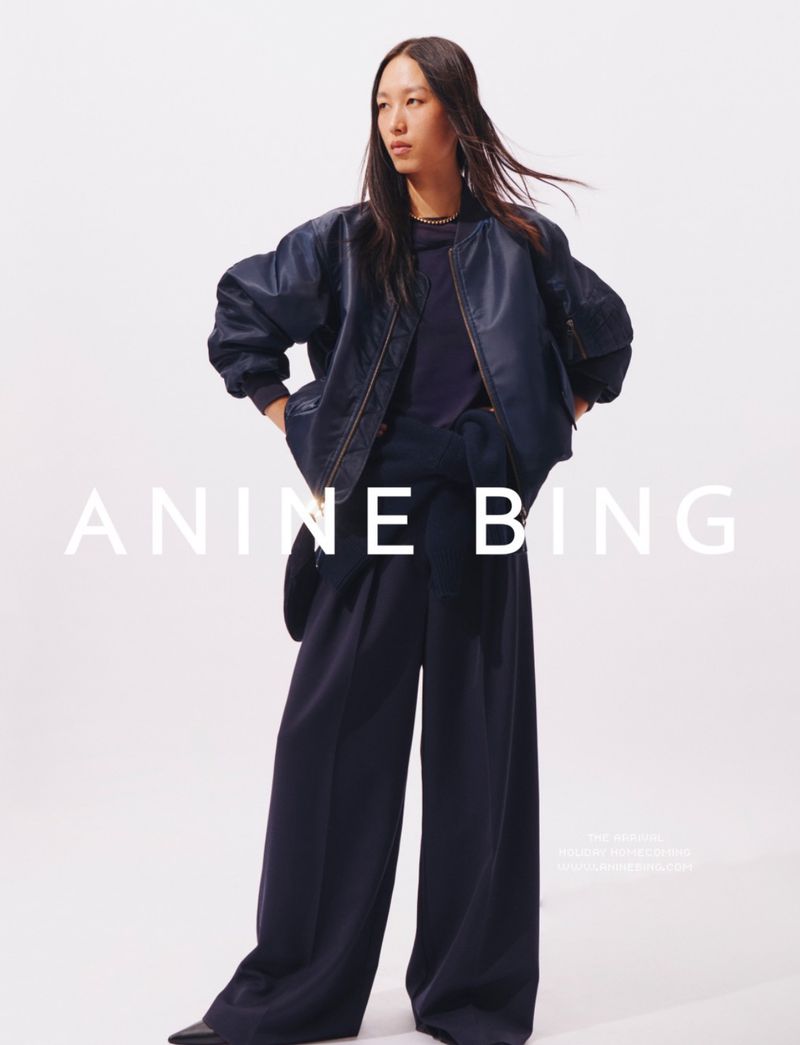 A stylish Asian woman in a dark blue bomber jacket and wide-leg pants poses against a bright white backdrop for a fashion editorial.