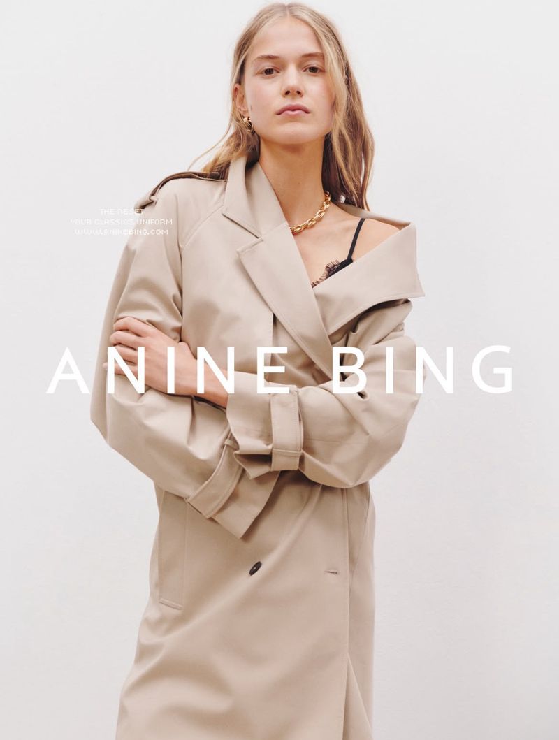 Fashion model in a trench coat poses indoors against a white background in a studio setting, showcasing modern apparel.