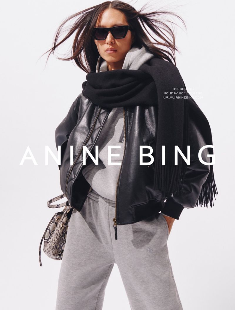 Fashion shot of a woman wearing sunglasses, leather jacket, and scarf, posing in a light studio. ANINE BING branding.