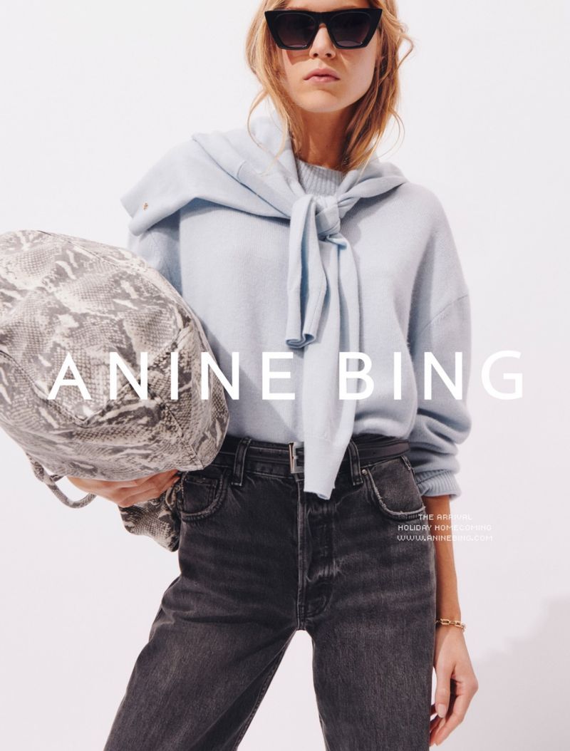 A fair-skinned woman in sunglasses, jeans, and sweater, poses in a studio with a snake-print purse and 'Anine Bing' logo.