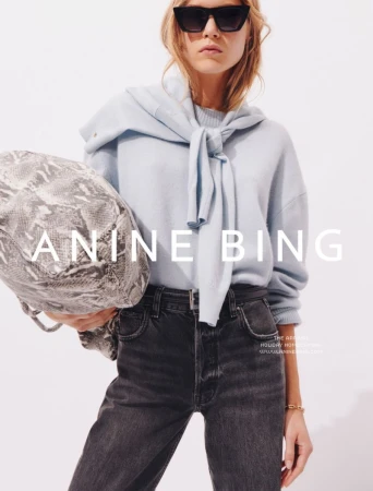 A fair-skinned woman in sunglasses, jeans, and sweater, poses in a studio with a snake-print purse and 'Anine Bing' logo.