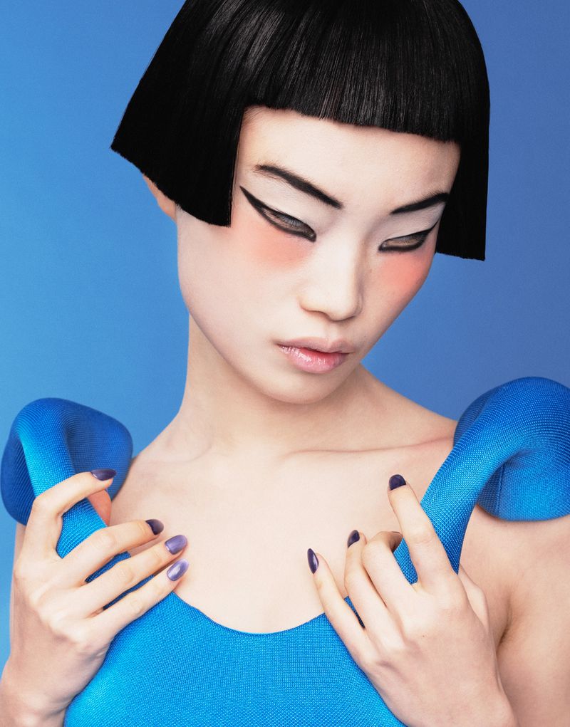 Asian woman with striking makeup, short bob hairstyle, and a blue textured top against a light blue background in a beauty style portrait.