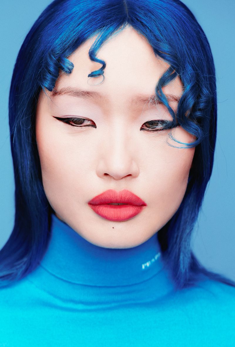 Close-up portrait of a woman with vibrant blue hair, dramatic winged eyeliner, and bold red lipstick against a soft blue backdrop.