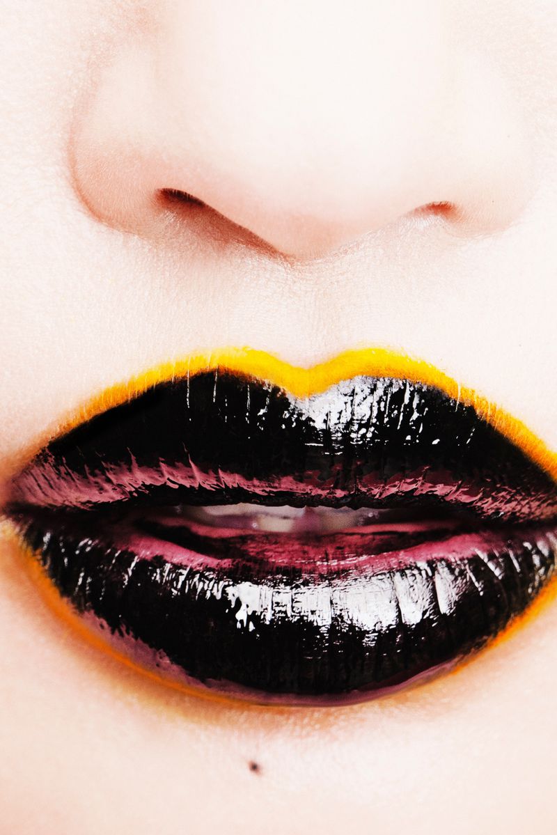 Close-up shot of a person's face featuring glossy black lipstick with a yellow outline on their lips. The image highlights beauty and style.