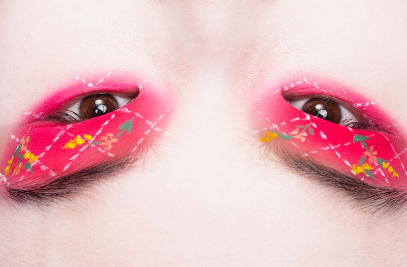 Extreme close-up of a face featuring vibrant pink eye makeup with delicate floral details and geometric line work on and around the eyelids.