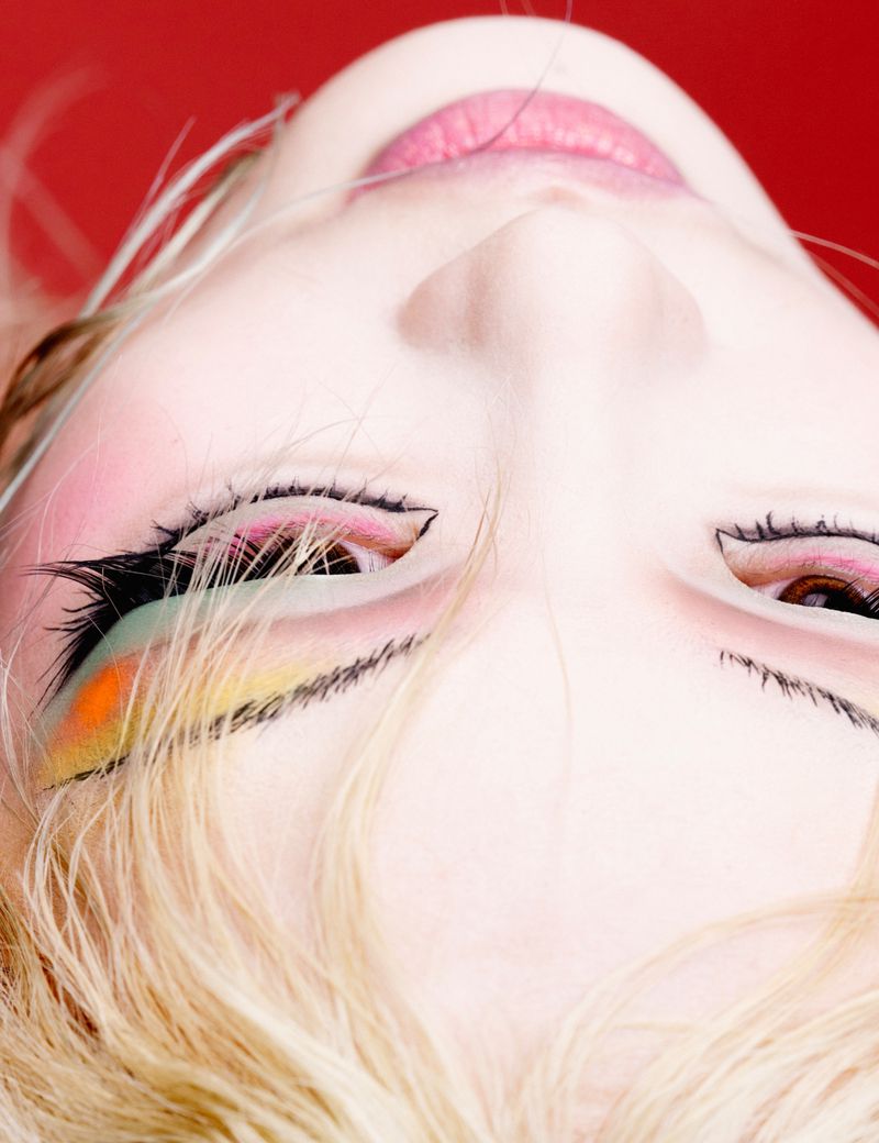 Extreme close-up of a person's face with creative makeup, featuring dramatic eyelashes and vibrant eyeshadow on a red background.