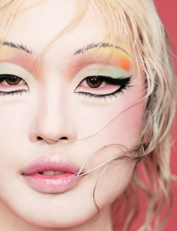 Close-up of a fair-skinned person with blond hair and dramatic eye makeup in peach, green, orange, and black with pink lips on a pink background.