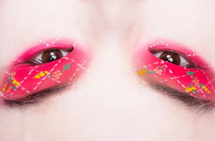 Close-up of a person's eyes featuring vibrant pink eye makeup with white stitched argyle patterns and floral details.