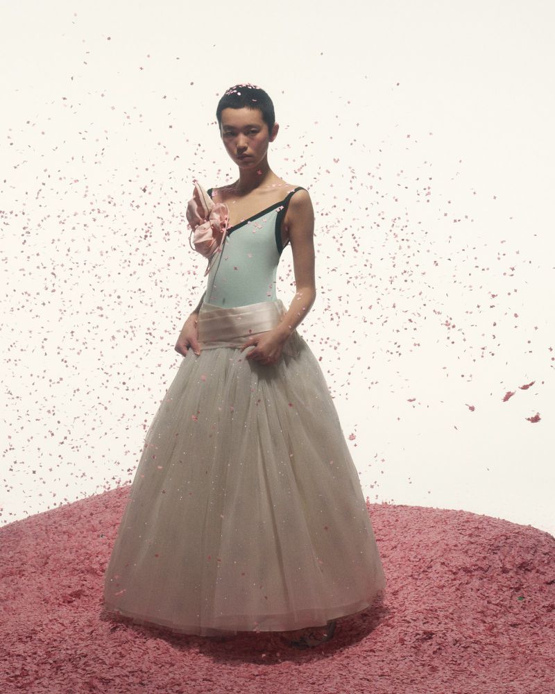 Elegant woman in light blue leotard and tulle skirt standing on a pink surface with confetti against a white backdrop.