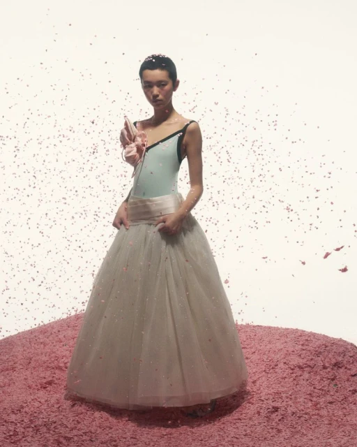 Elegant woman in light blue leotard and tulle skirt standing on a pink surface with confetti against a white backdrop.