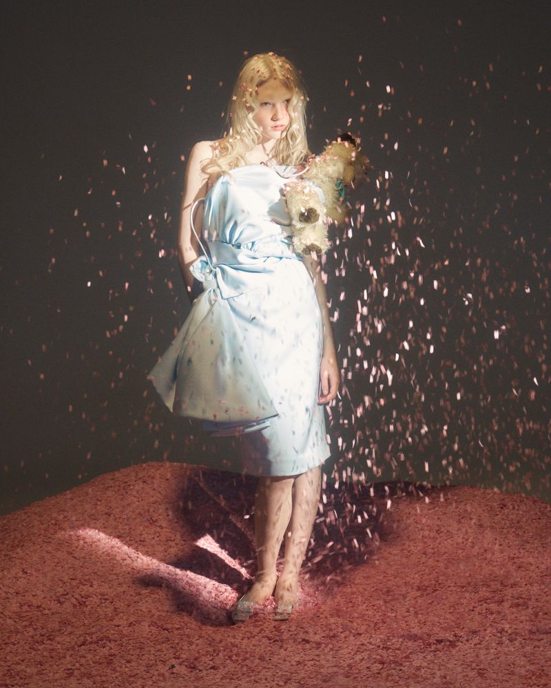 A young woman with blonde hair stands barefoot in a short, light blue dress, surrounded by shiny, reflective specks of light.
