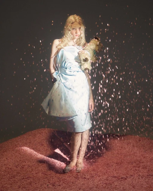 A young woman with blonde hair stands barefoot in a short, light blue dress, surrounded by shiny, reflective specks of light.