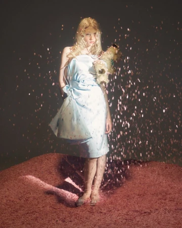 A young woman with blonde hair stands barefoot in a short, light blue dress, surrounded by shiny, reflective specks of light.