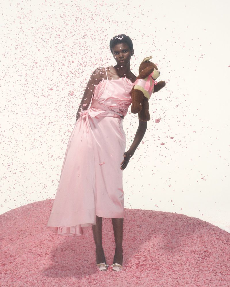 Fashion portrait of a Black female model in a pink dress, holding a teddy bear, standing in pink confetti with confetti falling around her.